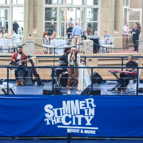 A band is performing on a stage with a sign that reads "Summer in the City". People are dining and socializing in the background.