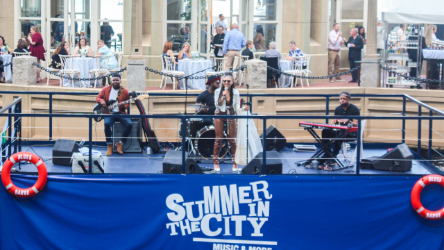 A band is performing on a stage with a sign that reads "Summer in the City". People are dining and socializing in the background.