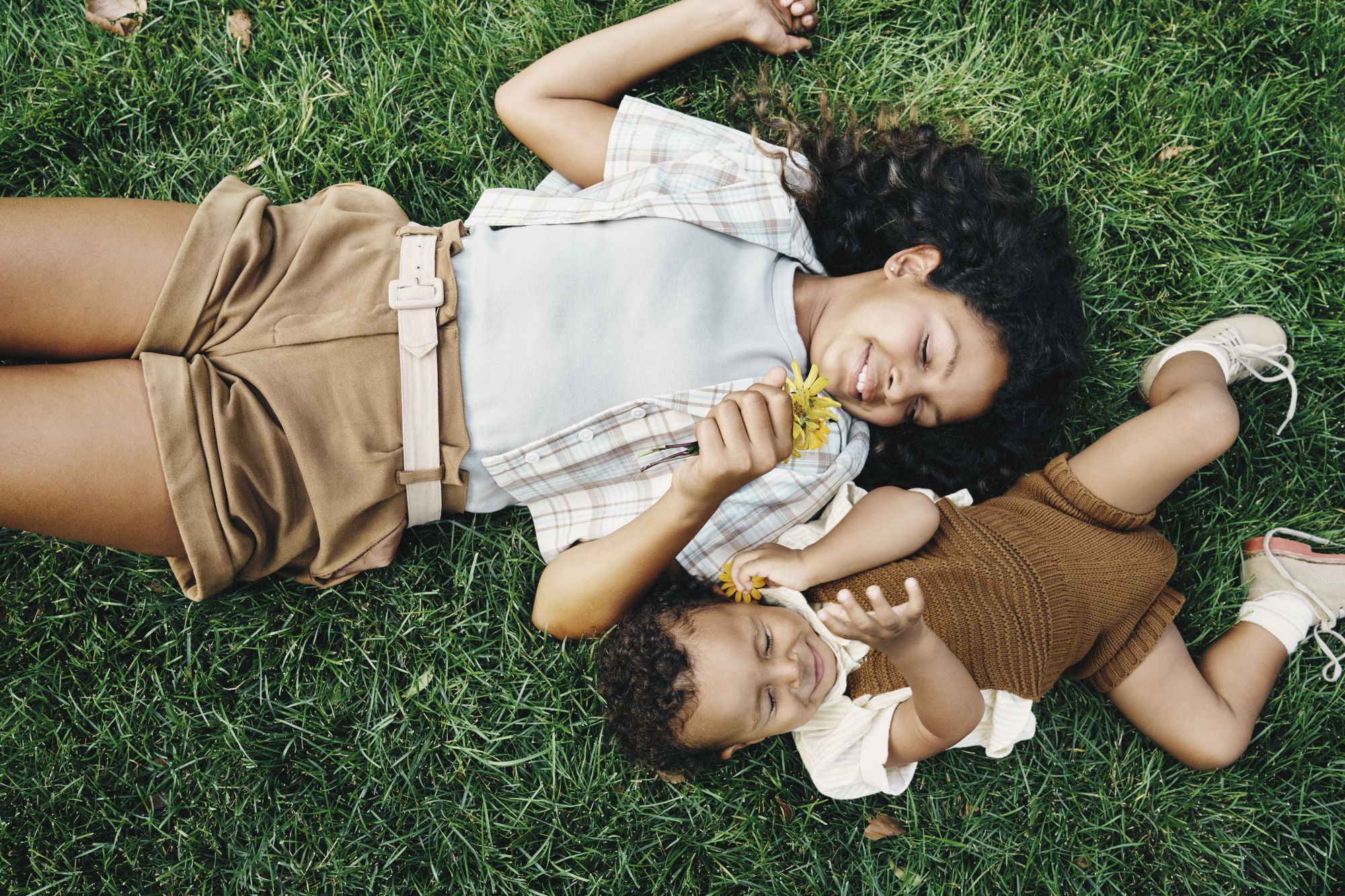 Two friends lie on grass, relaxing and sharing snacks on a sunny day, smiling as they chat and enjoy each other&rsquo;s company.