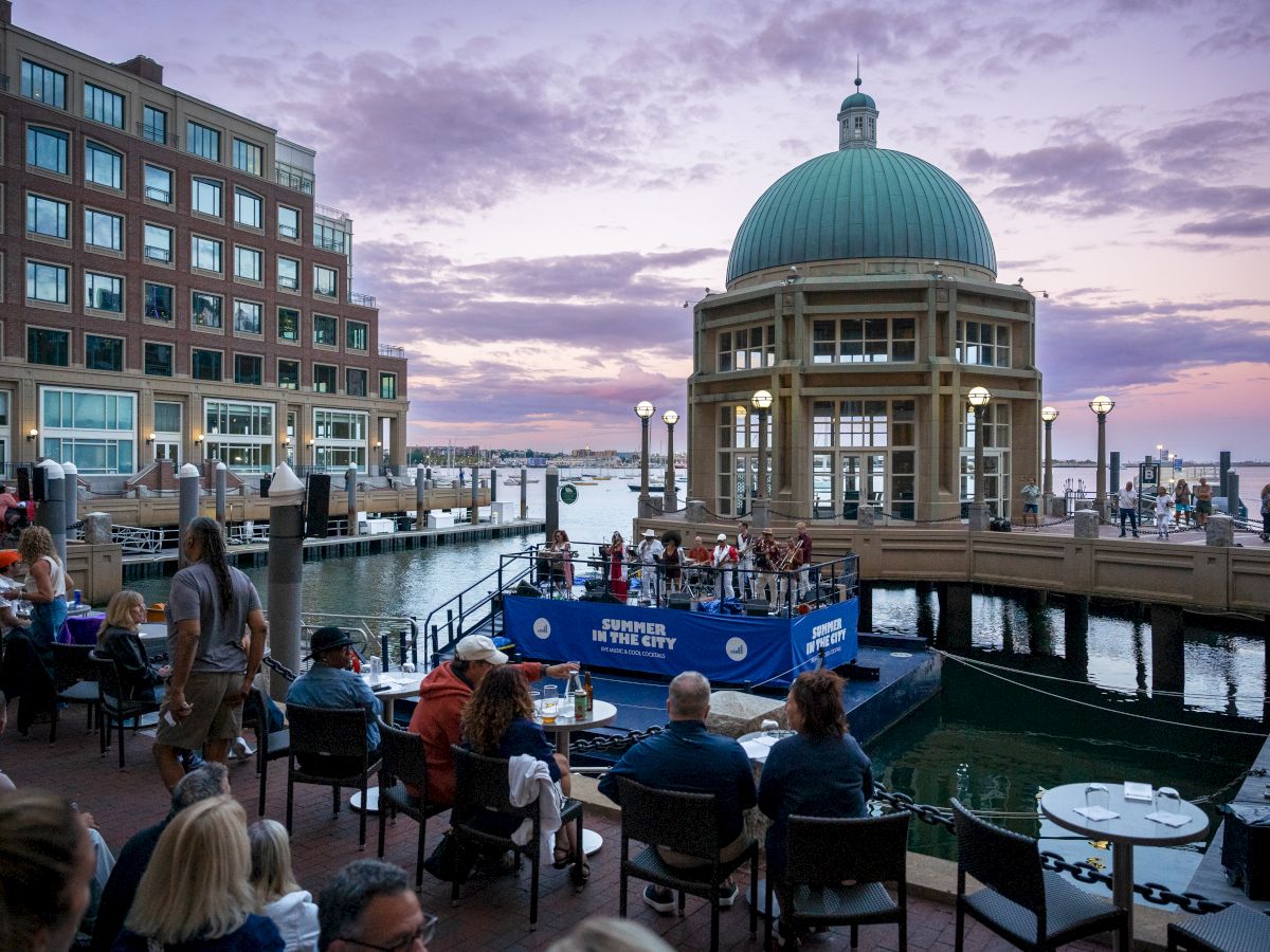 A waterfront venue with people watching a blue boat show near a domed pavilion at sunset, by the pier.