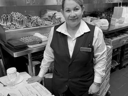 A person in a uniform stands beside a table set with dishes and utensils in a kitchen or food service area.