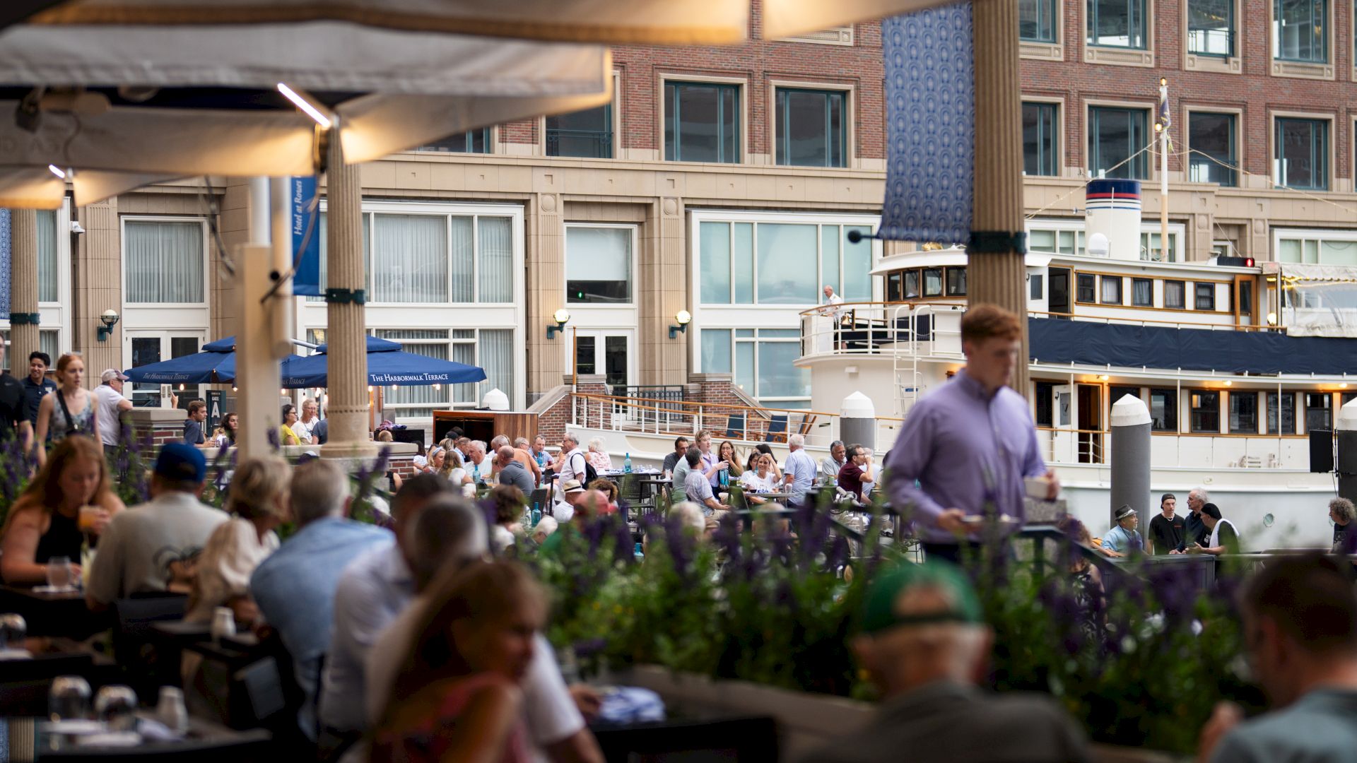 A vibrant waterfront scene with people dining outdoors, a docked boat, and a building with large windows in the background.