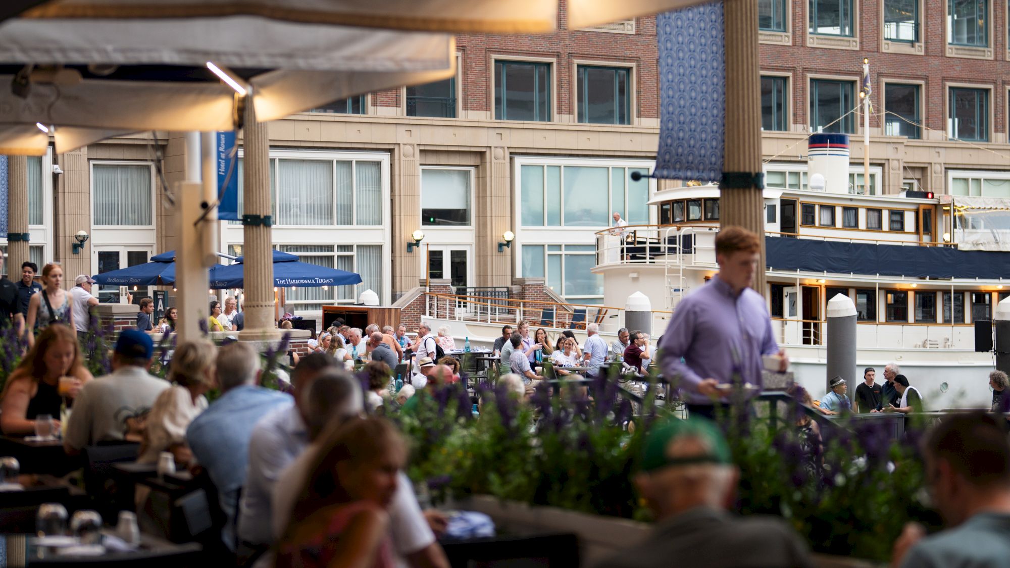 A vibrant waterfront scene with people dining outdoors, a docked boat, and a building with large windows in the background.