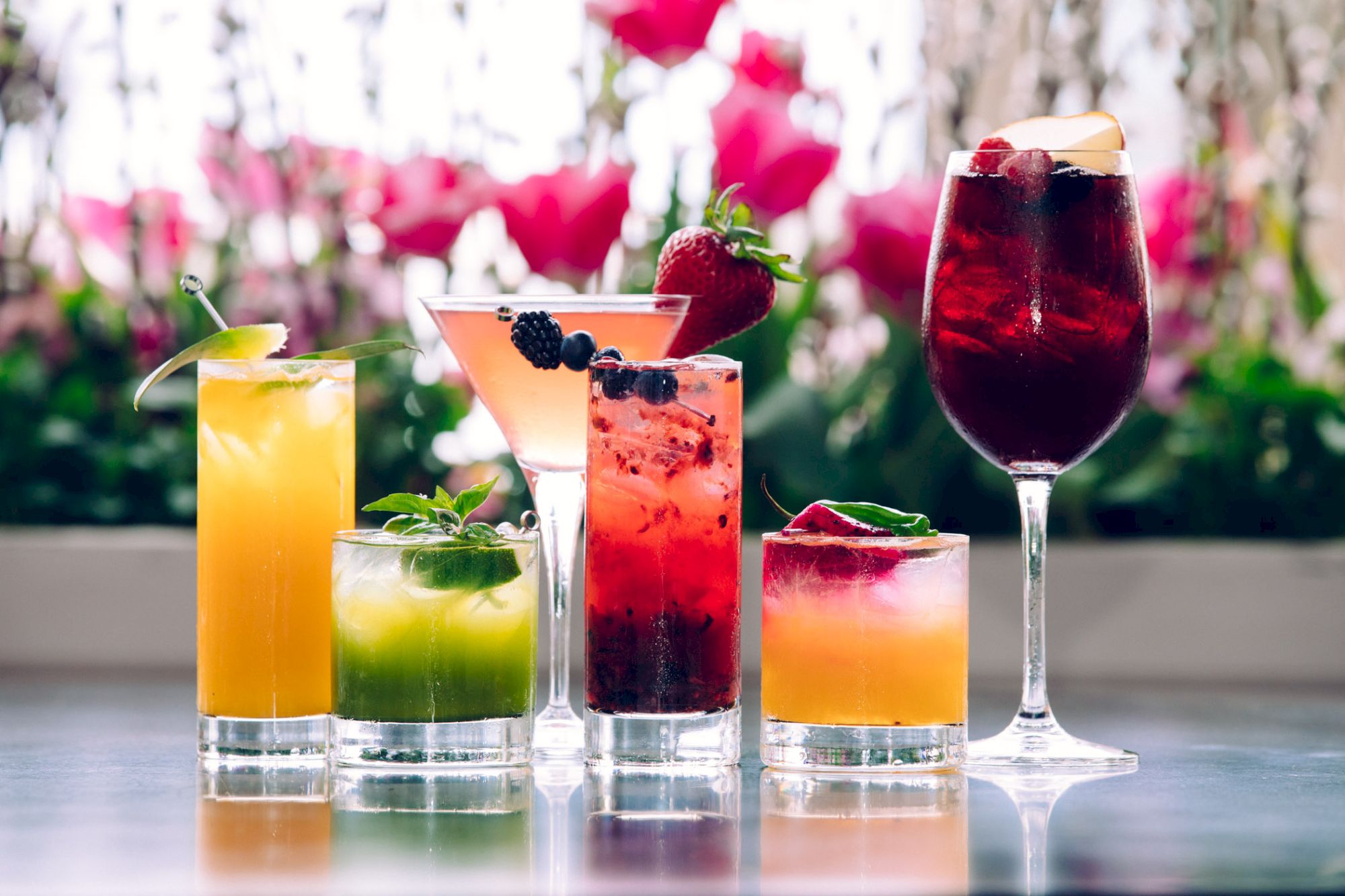 An array of colorful cocktails garnished with fruits and herbs, displayed against a blurred floral background.