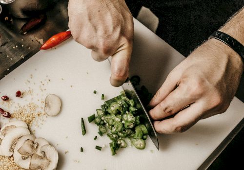 A person is chopping green vegetables on a cutting board. Nearby are sliced mushrooms, chopped meat, and various other ingredients.