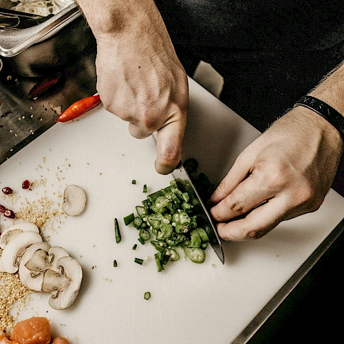 A person is chopping green vegetables on a cutting board. Nearby are sliced mushrooms, chopped meat, and various other ingredients.