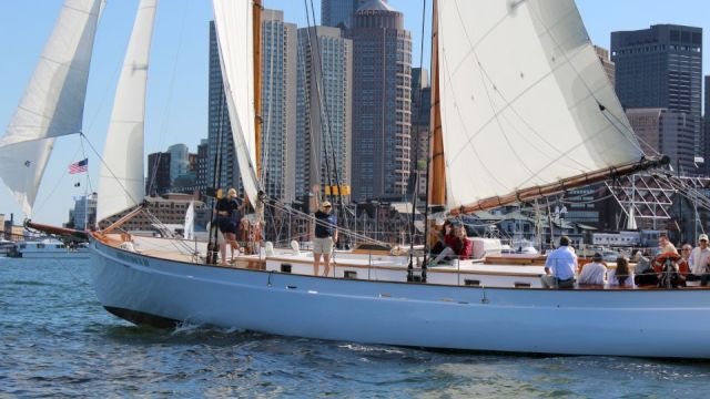 A sailboat with people onboard is sailing in the water, with a cityscape featuring tall buildings in the background.