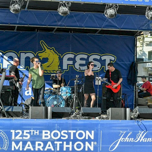 A band performs on stage at the 125th Boston Marathon with the backdrop reading "Fan Fest." John Hancock is one of the sponsors, as seen on signage.