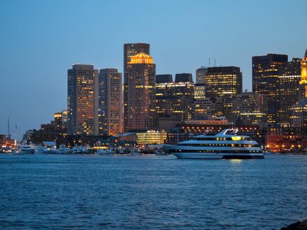 A city skyline at dusk with tall buildings illuminated and a large boat cruising on a body of water in the foreground.