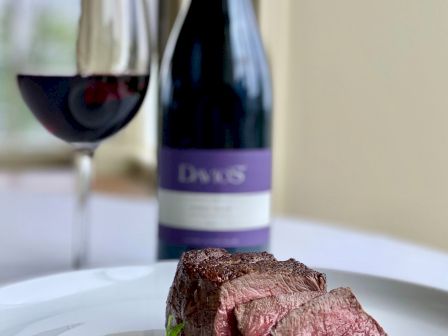 A plate with slices of steak, a garnish, a glass of red wine, and a bottle of wine in the background.