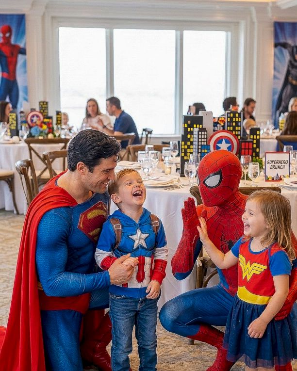 Three people dressed as superheroes (Spider-Man, Superman, Wonder Woman) pose with two kids at a party, smiling and chatting in a decorated room.