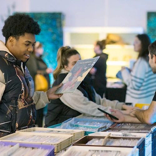 People browsing and chatting at a record store or market, with a focus on a person holding a vinyl record while another looks on interested.