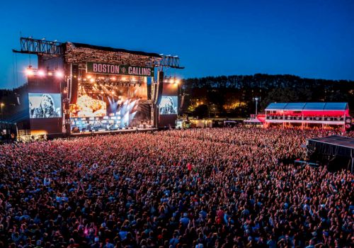 A large outdoor concert with a huge crowd and a stage lit up with colorful lights at night. The event is named "Boston Calling".
