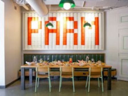 The image shows a dining area with a long wooden table and chairs. The wall has a large, brightly lit sign that reads "PARM," with two green lamps above.