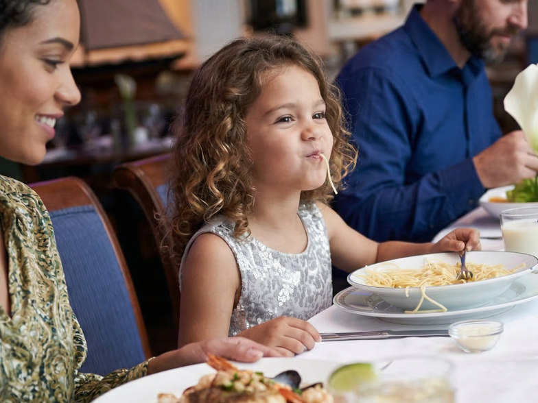 A family sits at a restaurant table enjoying a meal, with plates of food, drinks, and flowers on the table.