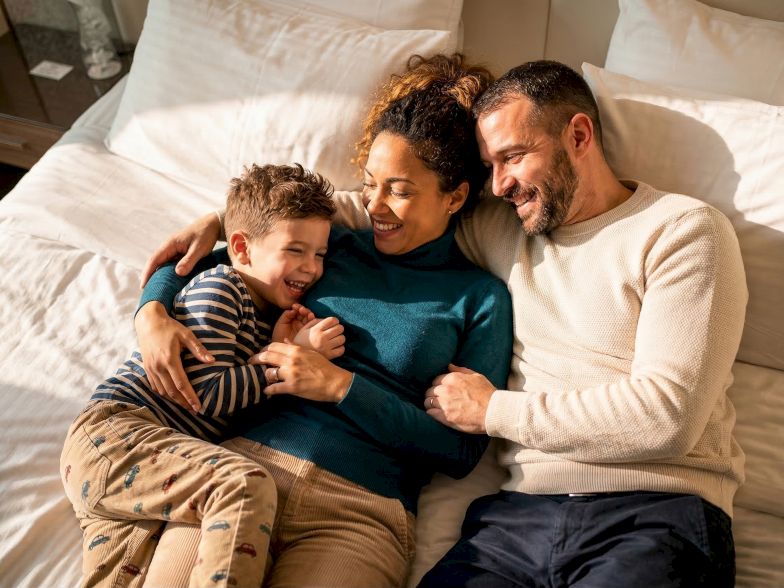 A family of three is laughing together on a bed, cuddling with the child between two adults, radiating warmth and joy.