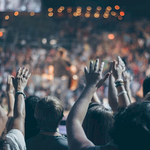 People are raising their hands in a crowded, lively event, likely a concert or a large gathering, with vibrant lights and a blurred background.