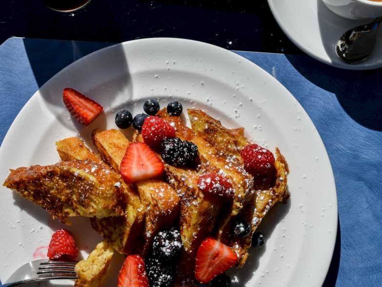 A plate with French toast topped with berries (strawberries, blackberries, raspberries) on a blue table, with a coffee cup nearby, sunny setting.