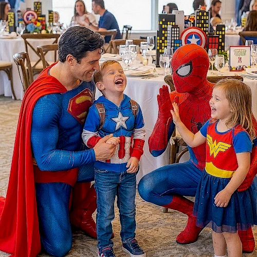Superhero-themed party: adults dressed as Superman and Spider-Man chat with two kids in capes and costumes, smiles and a festive table setting behind them.