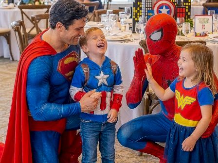 Superhero family moment: adults dressed as Spider-Man and Captain America cheer a laughing boy while a girl in a Wonder Woman outfit talks nearby, at a crowded party.