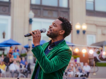 A man in a green jacket sings into a microphone outdoors with a crowd cheering behind him.