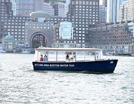 A blue water taxi labeled "Boston Water Taxi" is on the water in front of a cityscape with prominent buildings in the background.
