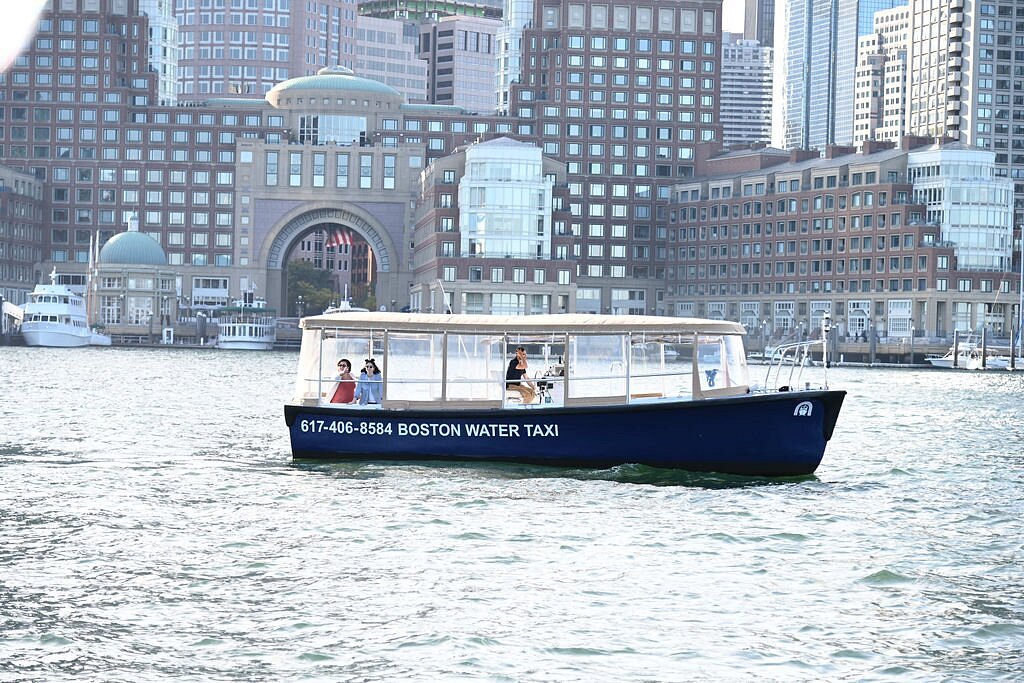 A blue water taxi labeled "Boston Water Taxi" is on the water in front of a cityscape with prominent buildings in the background.