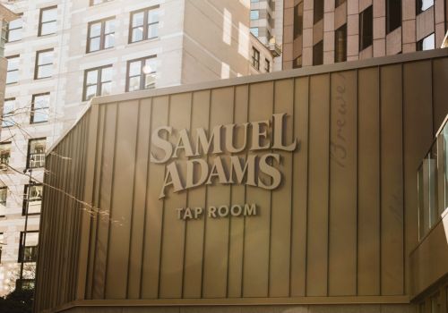 The image shows a building with the signage "Samuel Adams Tap Room," surrounded by tall buildings in an urban setting.
