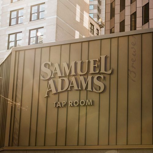 The image shows a building with the signage "Samuel Adams Tap Room," surrounded by tall buildings in an urban setting.