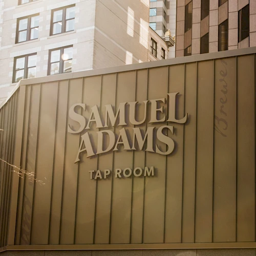 The image shows a building with the signage "Samuel Adams Tap Room," surrounded by tall buildings in an urban setting.