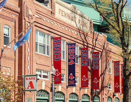 A brick stadium facade with banners showing years from 1903 to 198? hanging along the entrance, flagpoles, and a gate marked &ldquo;Gate A.&rdquo;