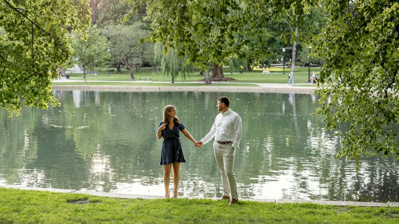 A couple stands by a pond in a park, holding hands and looking at each other under hanging tree branches.