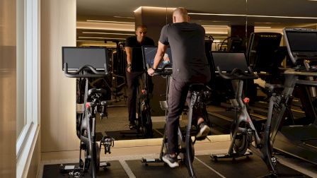 People working out on stationary bikes at a gym, facing a large mirror; warm lighting and modern cardio equipment visible.