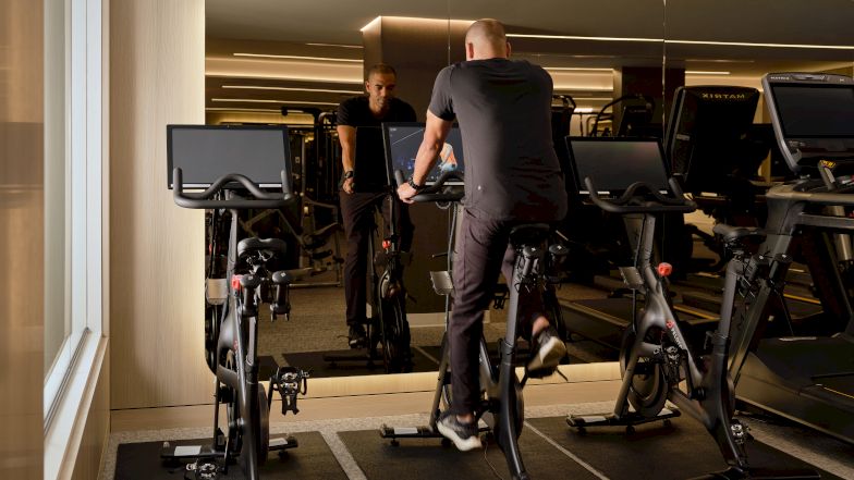 People working out on stationary bikes at a gym, facing a large mirror; warm lighting and modern cardio equipment visible.