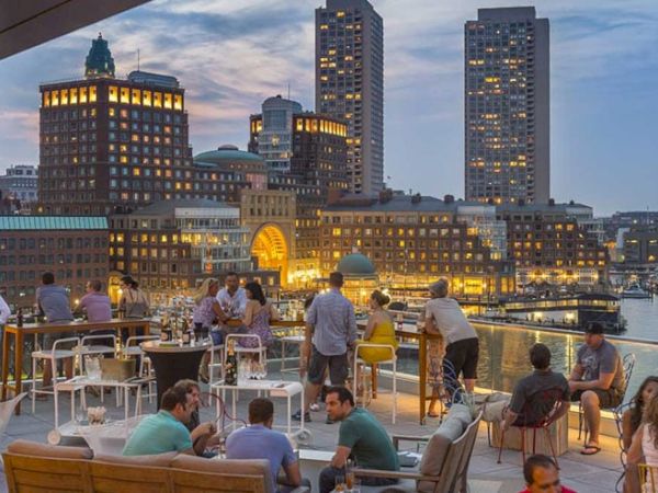 An evening at a rooftop bar with people socializing, overlooking a cityscape and waterfront.