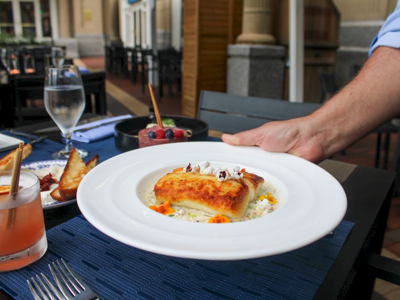 A plated dish of creamy, orange-tlecked lasagna or souffle with delicate garnishes sits on a white plate; a hand rests nearby at an outdoor table.