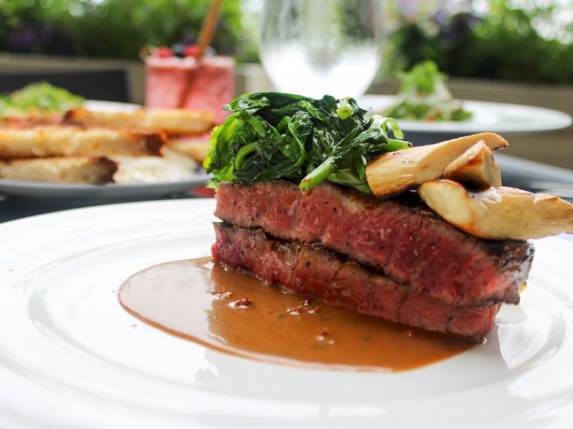A medium-rare beef steak on a plate, topped with greens, crispy pastry shards, and a rich brown sauce, with a blurred dish in background.
