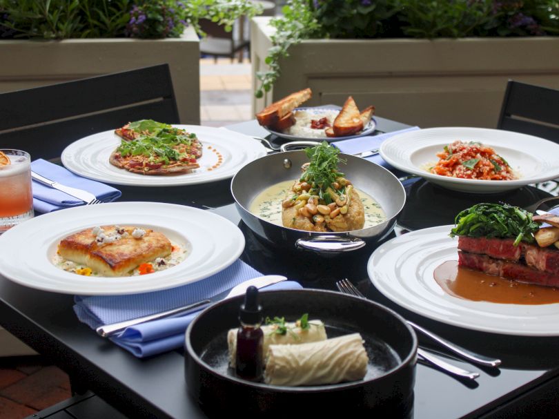 A variety of plated dishes on a table including salads, meat, pasta, a soup or sauce bowl, and drinks, all arranged beautifully for a meal.