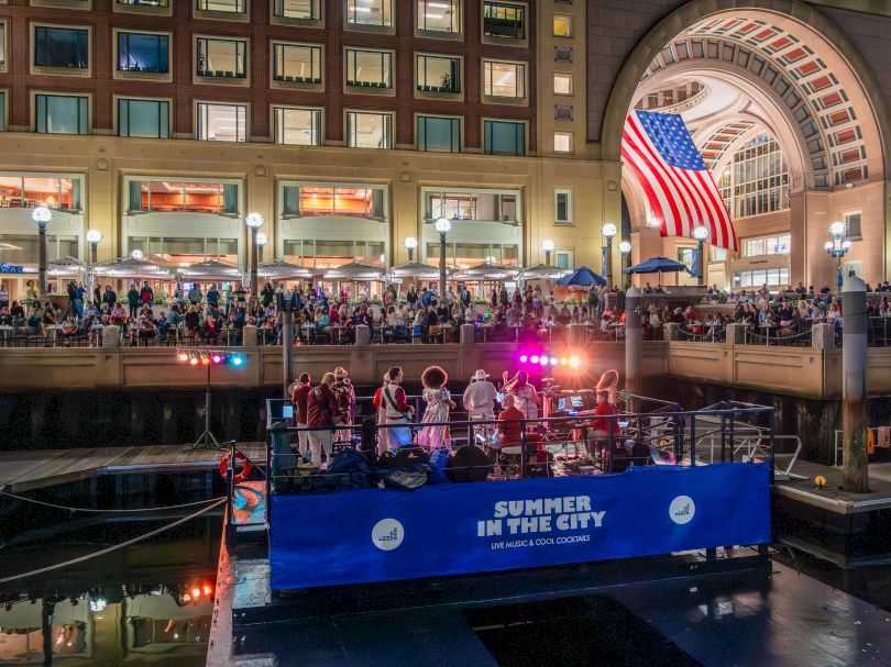 A lively waterfront scene at a city canal, with a blue party boat labeled &ldquo;Summer in the City&rdquo; docked near a grand arched bridge and a bustling crowd enjoying lights.