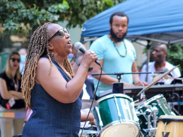 A woman is singing into a microphone with a band performing behind her. Others watch the performance outdoors under a canopy.