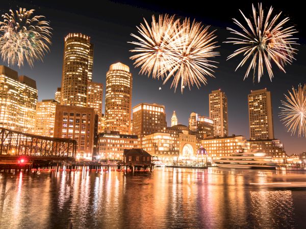 A cityscape at night with fireworks lighting up the sky, reflecting off the water. High-rise buildings and a bridge are prominently visible.