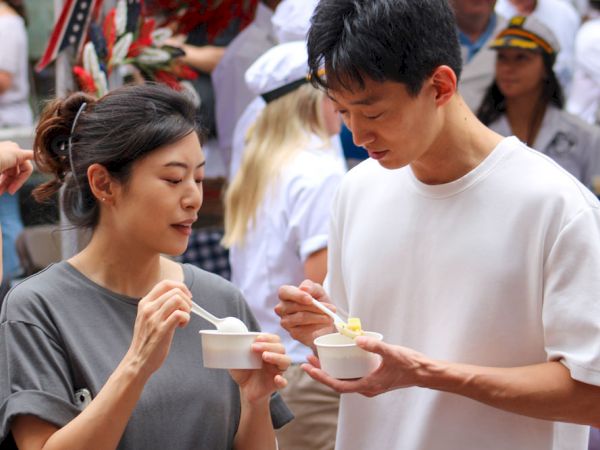 Two people enjoying food from small bowls, surrounded by others in what appears to be an outdoor event or market.