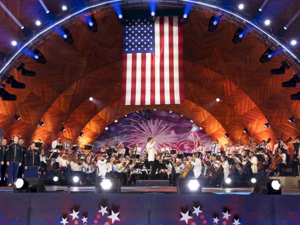 A large American flag hangs above a stage where an orchestra is performing. The venue is decorated with lights and star-themed decor, ending the sentence.