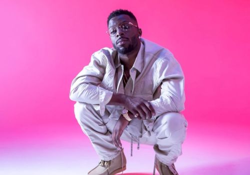 A man squats, wearing white clothes, against a pink backdrop.