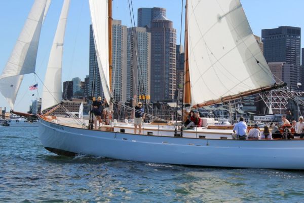 A sailboat with people onboard is sailing on the water, with a cityscape featuring tall buildings in the background.