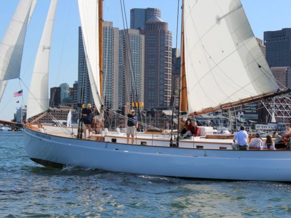 A sailboat with people onboard is sailing on the water, with a cityscape featuring tall buildings in the background.