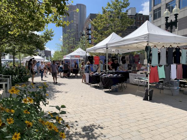 The image shows an outdoor market with several white tents, each selling different items such as clothes. People are walking around, and flowers are in the foreground.