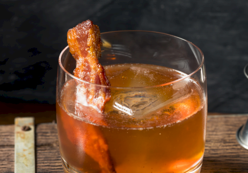 A cocktail garnished with a piece of crispy bacon, served in a glass with an ice cube, placed on a wooden surface.