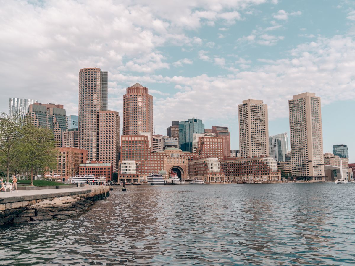 A city skyline with tall buildings along a waterfront, blue sky with scattered clouds, calm water in the foreground, and a pier on the left.