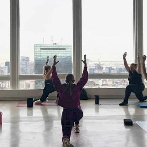 People doing yoga in a room with large windows, overlooking a cityscape. They are in a lunge pose with arms raised.
